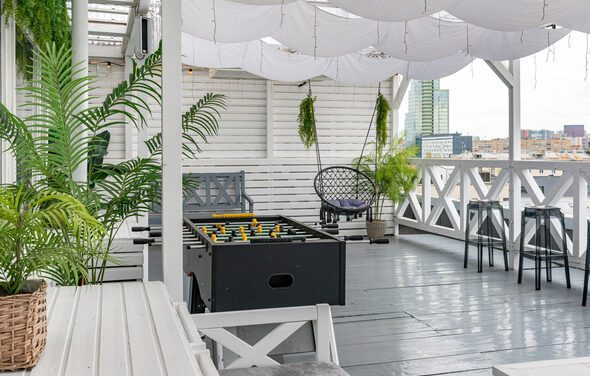 Лофт Май. Стильное пространство с верандой. Открытая терраса с серым деревянным полом, белыми стенами и тканевым потолком. Мебель: диваны, стулья, столики, футбольный стол. Растения в горшках. Вид на город.