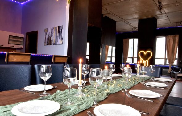 Элегантный лофт с хорошим звуком и балконом. Столовая зона с длинным деревянным столом, сервированным стеклянными бокалами и тарелками. В центре стола зеленая ткань и свечи. Мягкие стулья вдоль стола. На стене картина, сбоку кухня. Светодиодное освещение фиолетового цвета.