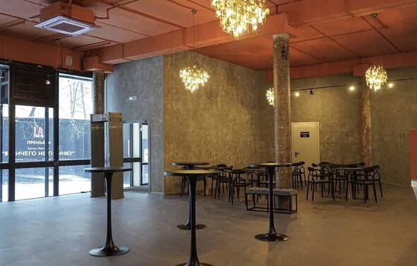 Театр в Хамовниках. Просторное помещение с высокими потолками, бетонные стены и пол, оранжевый потолок. Столики с высокими черными стойками и черными стульями. Большие окна, несколько круглых люстр. Прозрачные стеклянные двери, виден уличный баннер.