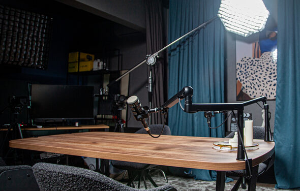 Студия для записи подкастов "Искусство Подкаста". Студия с деревянным столом, оборудованная микрофонами на стойках. Освещение достигается за счет большого софтбокса. На заднем плане шторы и декоративное панно. В помещении также есть стеллажи с техникой и экран.
