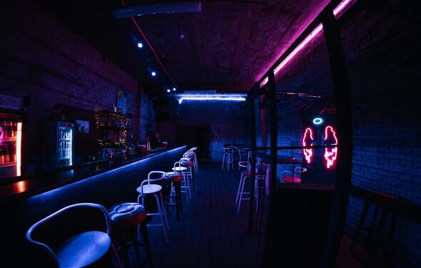 PVZ CLUB(ПВЗ Клуб). Бар с длинной стойкой, за которой стоят высокие стулья. Освещение неоновое, синие и розовые оттенки. На стене рисунок неонового ангельского крыла. Обстановка из тёмного кирпича, есть холодильник и полки с бутылками.