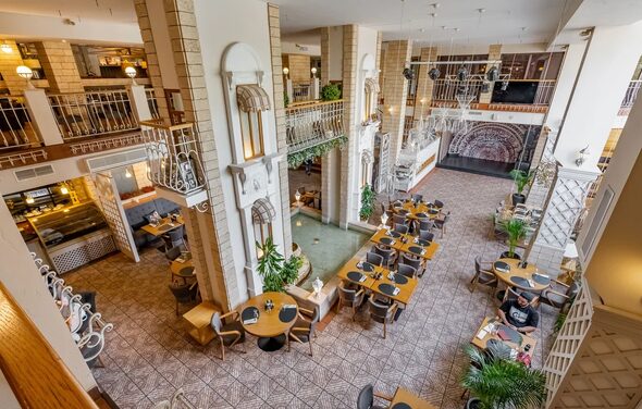 Ресторан Бульвар. Большое двухуровневое пространство с ресторанной зоной. Круглые деревянные столы с серыми стульями, плиточный пол. Балконы с решетчатыми перилами. Белые колонны и декоративные арки. Яркое освещение, живые растения.