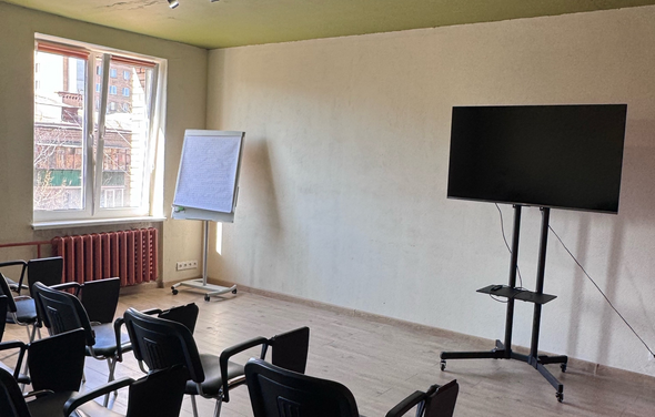 Первый План. Комната с деревянным полом, белыми стенами и зеленым потолком. Ряд черных стульев, флипчарт на треноге, телевизор на подставке. Большое окно, красный радиатор. Естественное освещение.