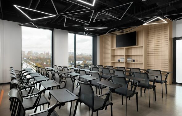 YES Technopark(ДА Технопарк). Конференц-зал. Конференц-зал с рядами черных стульев с пюпитрами, большие окна, современные геометрические светильники, полированные полы, деревянные панели на стене, экран с монитором, бутылки с водой на полке.