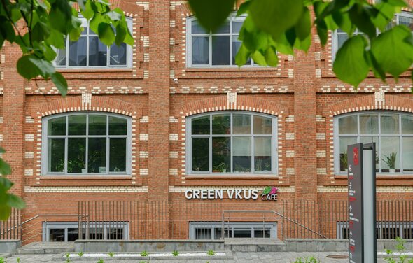 Green Loft(Зеленый Лофт). Фасад кирпичного здания с большими окнами в белой рамке. На уровне первого этажа вывеска "GREEN VKUS CAFE". Перед зданием находится металлическое ограждение и маленькая клумба. На переднем плане видны листья деревьев.