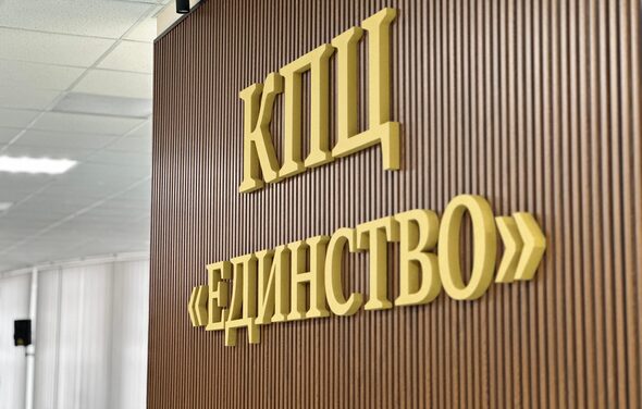 КПЦ Единство. Текст на стене: "КПЦ ЕДИНСТВО". Стена из вертикальных деревянных панелей коричневого цвета. Потолочные светильники с ровным мягким освещением. Белые потолочные панели.