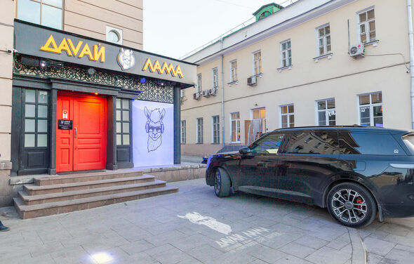 Давай Лама. Фасад здания с черной вывеской и оранжевыми буквами над красной входной дверью, изображение ламы на стене, ступени перед входом, припаркован черный внедорожник, окна здания на втором плане, серый асфальт, дневное освещение.