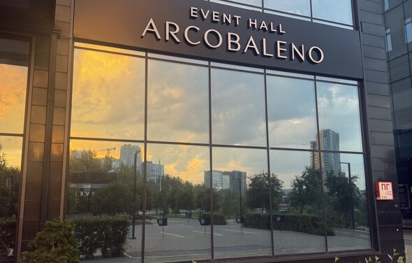 Event  Hall ARCOBALENO (Эвент Холл АРКОБАЛЕНО) . Фасад здания с большими стеклянными окнами, отражающими городской пейзаж с деревьями и небоскребами. Название на фасаде: «EVENT HALL ARCOBALENO». Вывеска «П» на стекле. Цвета: черный, стеклянные поверхности отражают закатное небо.