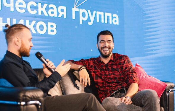 Конференц-зал до 150 гостей площадью 313 м². Современное пространство с ярко-синим фоном и текстом. Два человека сидят на кожаном диване. Один держит микрофон. На диване декоративные подушки розового цвета. Освещение мягкое, теплых тонов.