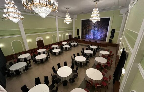 Банкетный зал Опера. Банкетный зал с высокими потолками и зелеными стенами, расставлены круглые столы с белыми скатертями и черными стульями. Вдоль стен стоят дополнительные стулья с узорчатыми спинками. Над сценой висят большие люстры.