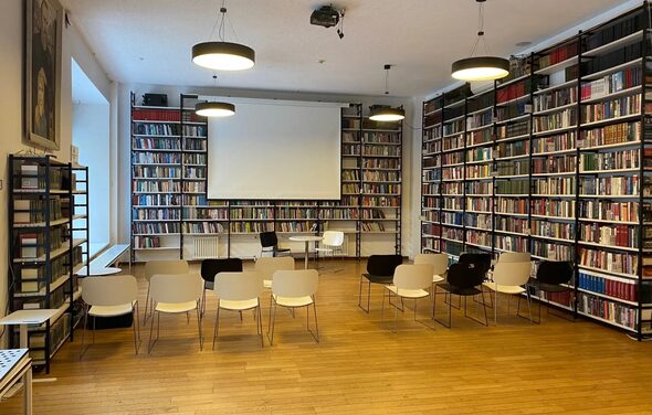 Зал для вашего мероприятия. Библиотека с деревянным полом и книжными полками вдоль стен. В центре установлены ряды стульев перед проектором и экраном. Освещение из подвесных ламп. На передней стене большие окна, справа — кресла и небольшой стол.