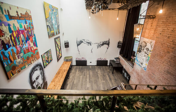 Street Art Loft (Стрит Арт Лофт). Лофт-пространство с высокими белыми стенами и кирпичной акцентной стеной. На стенах висят различные картины. Пол деревянный, из темных досок. Светильники подвешены к потолку. Помещение декорировано зеленью на ограждении.
