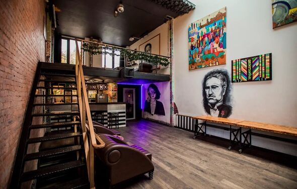 Street Art Loft (Стрит Арт Лофт). Лофт-пространство с высоким потолком, металлической лестницей и балконом. Стены украшены граффити и картинами. На полу темный ламинат. Коричневый диван у стены. Слева кирпичная кладка, справа белая стена с цветными картинами.