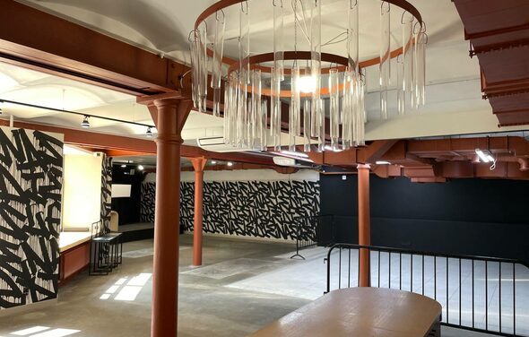 Бертгольд центр. Просторный лофт. Большое помещение с бетонным полом и высокими потолками. Красные металлические колонны, большие круглые светильники с декоративными элементами. Стены оформлены чёрно-белым узором. Присутствуют ограждения и мебель светло-коричневого цвета.
