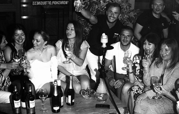 Work Shop Loft(Воркшоп Лофт). Зал 11 . Люди сидят вокруг низкого деревянного стола с напитками и кальяном. Справа на стене зелёные растения. В помещении полумрак, присутствуют бутылки и бокалы с напитками. На стекле надпись: "WORKSHOP LOFT BUSINESS".