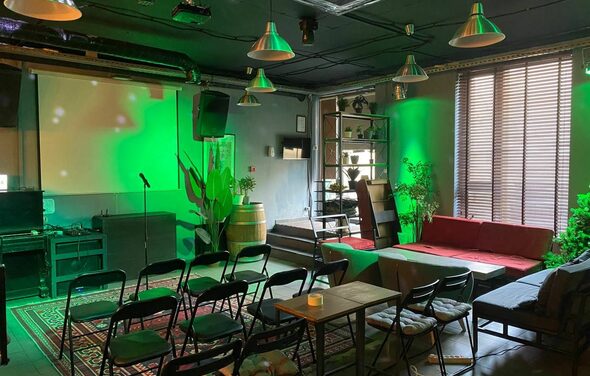 Work Shop Loft (Воркшоп Лофт). Зал 11 . Атмосферное пространство с черным потолком, подвесными лампами, зелёной подсветкой. Стулья в рядах, микрофонная стойка, проектор, экран, красный диван. Декор из растений, деревянные элементы, ковёр с узором, интегрированные полки, большие окна с жалюзи.