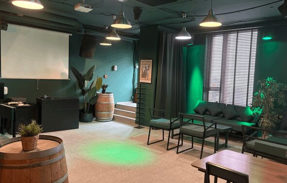 Work Shop Loft(Воркшоп Лофт). Зал 11 . Просторная комната с темно-зелеными стенами, большими окнами и жалюзи. Мебель: диваны, стулья, деревянные бочки, столы. Освещение представлено подвесными светильниками. На стене проекторный экран. В углу растения, постер на стене.