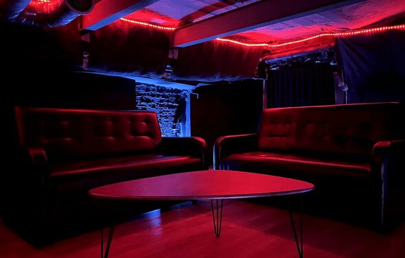 Лофт ИЗНАНКА. Зал ЛИХИЕ 90-е. Небольшое помещение с двумя кожаными диванами и круглым столом на металлических ножках. Потолок украшен красной светодиодной подсветкой. На стенах видна кирпичная кладка. Освещение приглушённое, цвета тёмные.