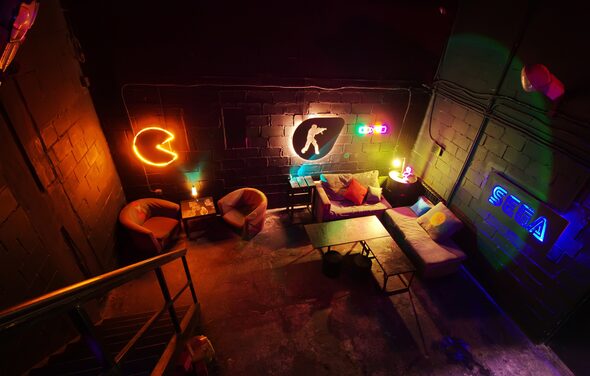 Лофт Геймеры. Тёмный лофт. Неоновые подсветки, игровые логотипы на кирпичной стене. Несколько кресел, столов, диванчиков. Фотография сделана со второго этажа