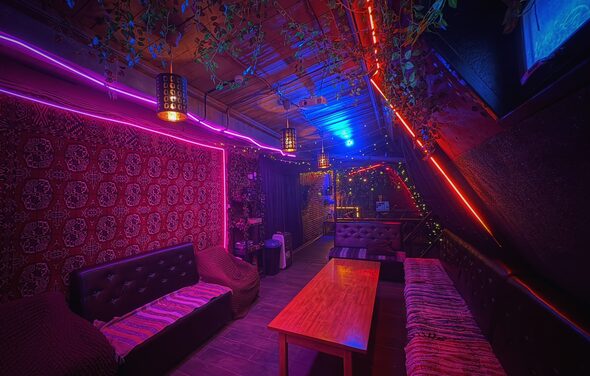 Лофт-бар. Клубное пространство с затенённой мебелью, включающей кожаные диваны и деревянный стол. Яркое неоновое освещение в синей и фиолетовой гамме. Декор из искусственных растений на потолке. На стенах тёмные панели и узорчатый ковёр.