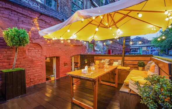 Cozy loft with bright accents . Открытая терраса с деревянным настилом и кирпичной стеной. Деревянный стол и скамейки с желтыми подушками. Над столом жёлтый зонт с гирляндой лампочек. Растения в горшках. Яркие граффити на стенах.