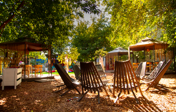 Лофт с территорией под открытым небом. Открытое пространство с деревянными шезлонгами на опилках, окружённое деревьями. Несколько беседок с занавесами и столами. Мягкое дневное освещение. Оттенки зелёного и коричневого.