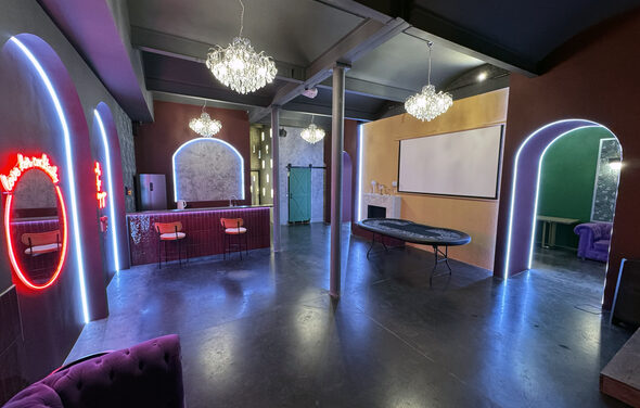 Белый лофт с яркой мебелью. Многофункциональное пространство с барной стойкой и высокими стульями, проектором на стене, стилизованными арками с неоновой подсветкой. Потолочные люстры, пол из темного материала, стены в теплых тонах.