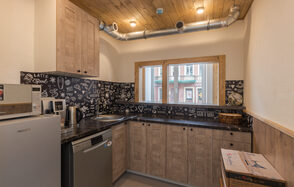 Светлый лофт с большими окнами. Кухня с деревянным потолком и светлыми стенами, мебель из светлого дерева, черные столешницы, черная плитка с белым рисунком, металлическая труба вентиляции, окно с видом на улицу, холодильник, посудомоечная машина, электрочайник.