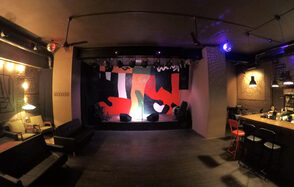 Лофт-бар со сценой. Сцена с ярким абстрактным фоном, оснащена светильниками. Справа барная стойка с высокими стульями. Мягкая мебель вдоль стен. Потолочные светильники и подвесные абажуры создают приглушенный свет. Красные и черные элементы декора.