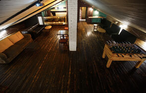 Duty Free Loft(Беспошлинный Лофт). Мансарда в центре Петербурга. Чердак с тёмным деревянным полом, кирпичной колонной, столами и мягкой мебелью. Поставлены диваны с подушками. В центре столы и стулья. На дальнем плане барная стойка. Есть настольный футбол. Освещение точечное и акцентированное.