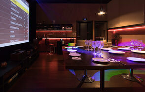 Караоке-лофт со светомузыкой. Темное помещение с большим столом, окруженным стульями. Посуда и бокалы аккуратно расставлены. На стене большой экран с меню. У стены барная стойка с высокими стульями и полками. Мягкое цветное освещение, деревянный пол, современные материалы.