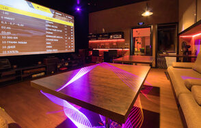 Караоке-лофт со светомузыкой. Пространство с деревянным столом и диванами, оснащённое большим экраном с интерфейсом меню. Современные светильники и разноцветное неоновое освещение. Деревянные полы, стены белые. Барная стойка с высокими стульями, коричневые и чёрные тона.