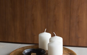 Светлый лофт со светомузыкой и барной стойкой. Три белые свечи на деревянной круглой подставке, рядом черная керамическая чаша с небольшими деревянными брусками. Фон из деревянных панелей теплого коричневого оттенка. Поверхность белая, гладкая.