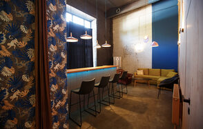 Дизайнерский лофт с высокими потолками. Барная зона с высоким столом и четырьмя черными стульями на фоне кирпичной стены. Справа светло-зеленый диван и кресло на фоне синей стены. Потолочные светильники над баром. Плотные шторы на окне. Текстиль с растительным узором.