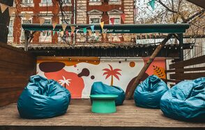 Лофт Surf (Серф). Тропический лофт с большой верандой.. Открытая площадка с деревянным полом, яркие пуфы в виде кресел, круглый стол, разноцветные флажки натянуты сверху, граффити с абстрактными узорами и пальмами на стене, забор из дерева, элементы зелени.