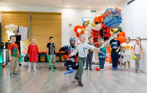 Лофт для ярких душевных вечеринок. Пространство с деревянным полом и белыми стенами. Центральное освещение, справа проекционный экран. Слева рольставни на окнах. Декорировано красными, черными и золотыми воздушными шарами. В помещении дети играют в костюмах, персонаж в костюме супергероя.