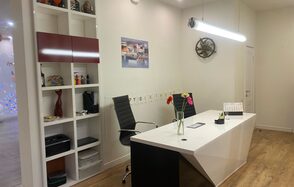 Лофт для ярких душевных вечеринок. Рабочее пространство с современным белым столом, за которым стоит черное офисное кресло. Справа стена с полкой, где размещены книги и украшения. На столе ваза с цветами и канцелярия. Светлое помещение с деревянным полом и подвесным освещением.