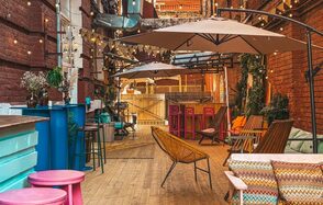 Двухуровневый лофт в гавайском стиле с кухней. Уличное пространство с деревянным полом и кирпичными стенами. Парасолей, светящиеся гирлянды, разноцветные барные стойки и стулья, кресла, деревянные и металлические табуреты. Растения в горшках, разнообразные узоры, подушки.