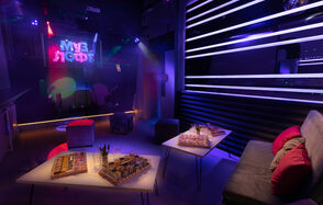 Музыкальный лофт на Тульской. Караоке-зал с неоновыми вывесками, фиолетовым освещением, диванами с красными подушками, квадратными столами с угощениями. Стены темные с горизонтальными световыми панелями. На заднем плане сцена с микрофоном. Надпись на неоне: "Муз Лофт".