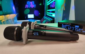 Музыкальный лофт на Тульской. Два беспроводных микрофона на деревянном столе, рядом цифровой приемник с синим дисплеем. На заднем плане разноцветные светодиодные элементы и мягкие диваны, яркие декоративные световые панели на стенах, тусклое разноцветное освещение.