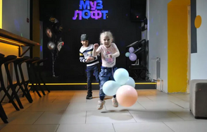 Музыкальный лофт на Тульской. Праздничное пространство с черной стеной и надписью "музлофт". Справа диван, слева высокие стулья у барной стойки. Плиточный пол. На полу разноцветные шарики. Освещение приглушенное, на стенах световые акценты.