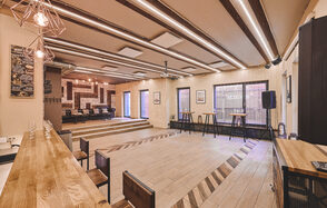 Loft Lodge 6. На фото показано просторное помещение с деревянным полом и ступенчатым подиумом. В центре сцена с диванами. Оборудование: нижние столы, стулья, барная стойка, динамик. Оформление: кирпичная стена, потолочные светильники. Цвета теплые, состояние хорошее.