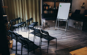 Лофт с возможностью зонирования. Небольшое помещение с деревянным полом, черные стулья в два ряда, флипчарт на треноге. В углу книжные полки и диваны с подушками. Тяжелые шторы на окне. Стены светло-серые, с декоративными вставками. Освещение мягкое.