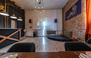 Rubin Loft (Рубин Лофт). Зал Джаз. Лофтовое помещение с кирпичной стеной, проектор на стене, сцена, стулья, украшенные текстилем; две люстры, барная стойка с деревянной отделкой и три подвесных светильника. Пол светлый, справа красные шторы.