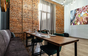 Rubin Loft (Рубин Лофт). Лофт средний. Лофт-пространство с кирпичными и белыми стенами, деревянный стол, стулья с тёмной обивкой, длинный мягкий диван, подвесное освещение, большая фотография на стене, высокий потолок, деревянный пол, окно с занавесками.