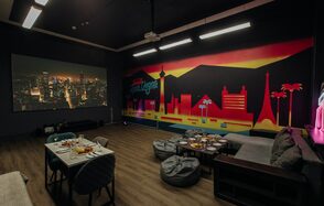 Зал Лас-вегас. Лофт-зал в американском стиле . Комната с проектором и большим экраном, диванами, пуфами и столом с сервировкой для еды. На стене яркое изображение городского пейзажа с надписью. Темное освещение, светлые деревянные полы, подвесные светильники.