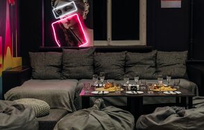 Зал Лас-вегас. Лофт-зал в американском стиле . Большая комната с темными стенами, серые диваны с подушками, низкий стол с тарелками и стаканами. Неоновая вывеска в форме снимка человека, светлое окно, несколько кресел, мягкое освещение.