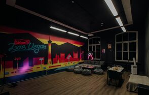 Зал Лас-вегас. Лофт-зал в американском стиле . Темное помещение с граффити на стене, изображающем ночной Лас-Вегас. Пол деревянный, потолочные светильники продолговатые. В углу зона с серыми пуфами и столиком. Под неоновым знаком стоят кушетка и стул. Большие окна без штор.