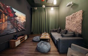 Зал Китай. Лофт-зал в китайском стиле . Компактное помещение с современным интерьером. Мягкий серый диван, пуфы, деревянный стол. Большой экран на стене, ковровое панно. Потолочные светильники, зелёные шторы. Напольное покрытие из дерева. Белый проектор на потолке.