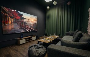 Зал Китай. Лофт-зал в китайском стиле . Кинотеатр с серым диваном и пуфом, деревянный пол, на стене большой экран с изображением пейзажа. Зеленые шторы на окне, на потолке подвесные светильники. Перед диваном низкий столик с сервировкой бокалами и тарелками.